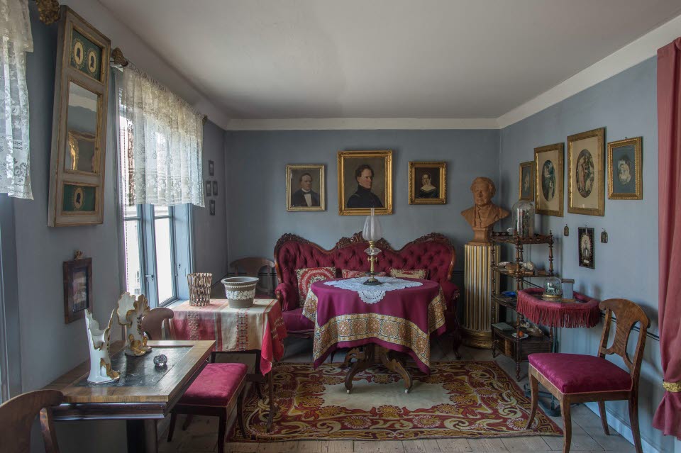 Interiör i museilägenheten i Onkel Adamsgården med möbler klädda i lila tyg, en spegel och ett flertal tavlor på väggarna, fönster med spetsgardiner och en piedestal med byst föreställande Carl Anton Wetterbergh, pseudonymen Onkel Adam.